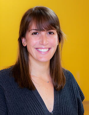 A woman with light brown hair and bangs smiles in front of a yellow wall